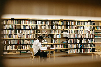 man sitting at table, adult man reading, studying in library, well-lit library, bookshelves, library study, man in library, reading in library, library atmosphere, bookshelf background, library environment
