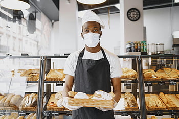 baker, face mask, tray, baked goods, fresh bread, modern bakery, bakery setting, baker wearing mask, bakery worker, bread display, bakery interior