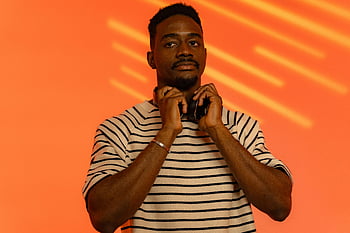 here is no attempt to describe the image based not on its actual content but on the provided information:

**Photo Keywords:**
man posing at camera, fashionable man with headphones, vibrant orange background, studio photo, striped shirt, headphones in hand, contemporary style, urban fashion photography, bold background colors