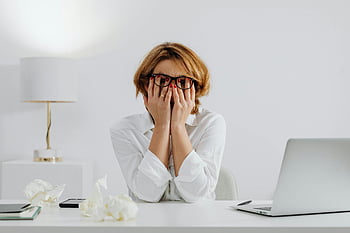 woman, white long sleeve shirt, eyeglasses, sitting at table, desk, laptop, tissues, stressed, hands on face, office setting, work stress