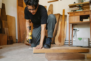 ethnischer Mann, Loch bohren, Holzbrett, selbstbewusster Handwerker, Gut ausgestattete Workshop, Bohrmaschine, Holzbearbeitung, Werkstattumgebung, fokussierte Arbeit, Zimmermannswerkzeuge, ethnischer Handwerker