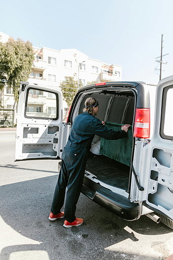 man in coverall, blue coveralls, standing at car, loading van, sunny day, moving service, worker, van, back of car, outdoor, urban setting
