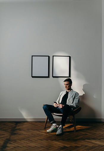 Mand i hvid kjole shirt, Sidder på sort stol, Brug af smartphone, Minimalistisk hjemmekontor, indrammet kunst, trægulve, indendørs indstilling, afslappet tøj, moderne interiør, afslappet kropsholdning, hjem indretning