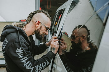 Mann mit Dreadlocks, Arbeiten am Autofenster, Tönung von Fahrzeugfenstern, fokussierter Mann, präzise Arbeit, Autofenstertönung, Fahrzeugtönung, Mann mit Brille, Mann mit Bart, Autofenster, Fahrzeugfenster