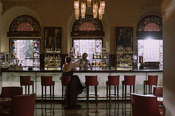 woman in black dress, bar counter, bartender conversation, stylish bar, luxurious bar, stained glass windows, bar interior, elegant setting, bar stools, chandelier lighting, bar atmosphere