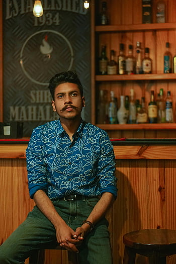 man sitting, brown wooden stool, wooden bar, array of spirits, fashionable young man, portrait, seated, building interior, stylish outfit, bar setting, spirits collection