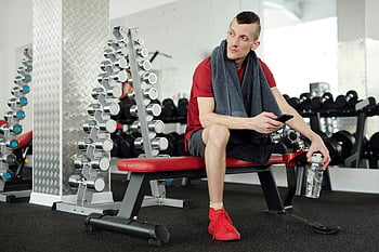 man i röd skjorta, svarta shorts, röd och svart stol, protesben, Gym, smartphone, vattenflaska, tar en rast, träna, kondition, handduk