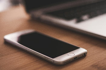 Grey Apple iPhone, smartphone, wooden table, blurred laptop, cozy indoor setting, tech devices, mobile phone, desk setup, technology, office environment, close-up shot