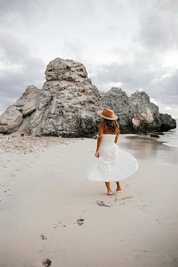 woman in white dress, beach walk, serene beach, back view, rocks background, white dress, beach scene, woman walking, beach rocks, beach fashion, hat and dress