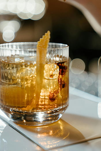 clear glass cup, yellow liquid, whiskey glass, citrus garnish, bar table, cocktail themes, beverage themes, close-up, ice cubes, orange peel, drinks