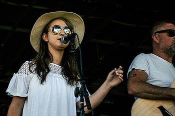 en haut blanc à manches courtes, concert en plein air, chanteuse, guitariste, performance sur scène, concert vibrant, microphone, des lunettes de soleil, chapeau de paille, guitare acoustique, performance passionnée