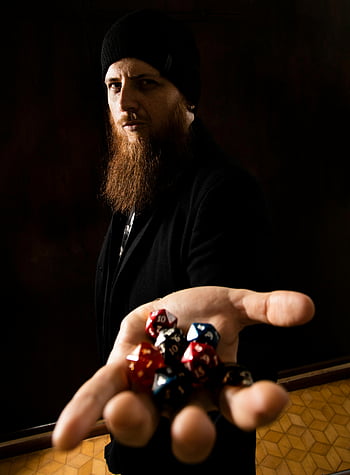 man with beard, holding dice, colorful dice, dark environment, dramatic lighting, bearded man, presenting dice, dark background, multi-colored dice, dramatic scene, black beanie