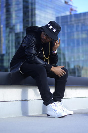 man sitting, glass window, urban setting, black jacket, hat, white sneakers, stylish outfit, cityscape background, modern architecture, casual fashion, street style