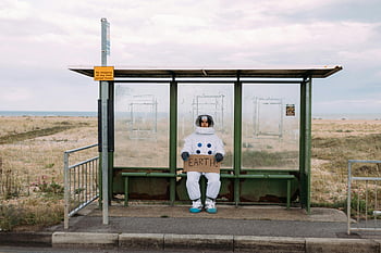 astronaut, space suit, rural bus stop, sign, Earth, alone, rural setting, bus stop, waiting, space theme, humorous scene