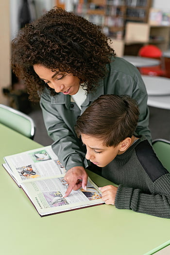 woman teaching, boy reading, classroom, green table, teacher helping, student learning, book, education, reading skills, literacy, teaching methods