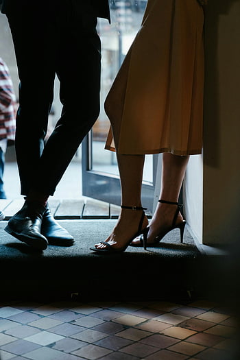 Žena v hnědých šatech, černé kožené boty, Stylová obuv, matně osvětlené nastavení, Pár u dveří, Elegantní pozice, vysoké podpatky, formální oblečení, vnitřní scéna, módní fotografie, Pár styl