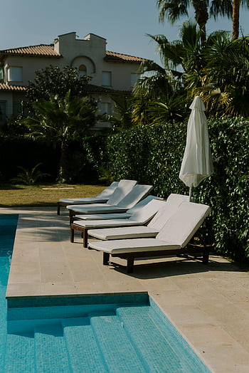lounge chairs, swimming pool, sunlit poolside, cushioned lounge chairs, lush greenery, summer retreat, poolside chairs, outdoor relaxation, poolside umbrellas, tropical setting, villa poolside