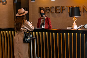 Wanita saling memandang, resepsi hotel, resepsionis mengenakan topeng, wanita berbicara, lobi hotel, topeng wajah, pelayanan pelanggan, perjalanan, bagasi, Pakaian Profesional, keramahan