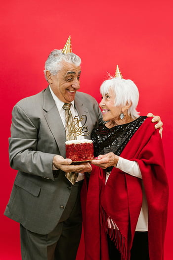 elderly couple, 50th anniversary, red cake, vibrant background, happy celebration, party hats, senior couple, anniversary cake, red background, festive occasion, elderly love