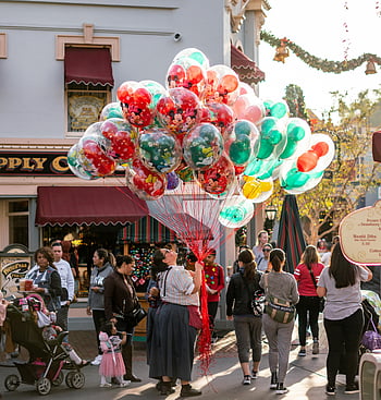 woman holding balloons, street scene, vendor selling balloons, bustling crowd, daytime festivities, vibrant balloons, lively street, people at the road, festive atmosphere, outdoor event, Disney characters