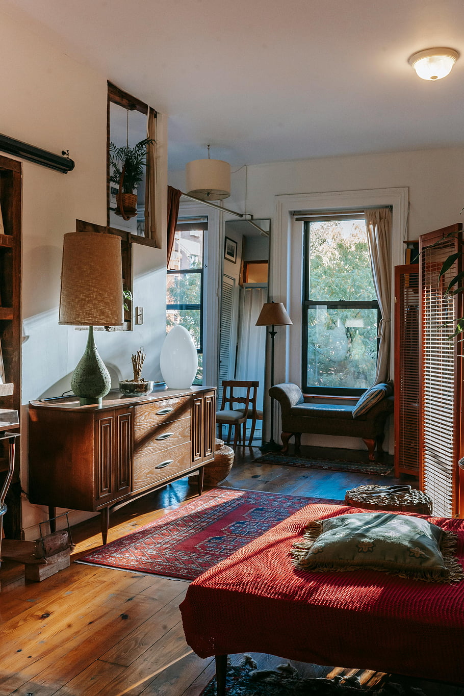 cozy bedroom, antique wooden furniture, stylish decorative elements, warm vintage-style, elegant decor, natural sunlight, wooden floor, red rug, wooden dresser, hanging plants, floor lamp