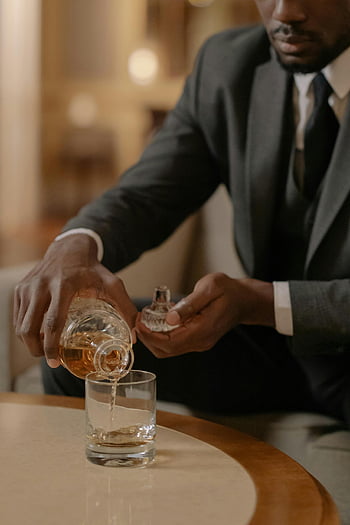 man in black suit, clear drinking glass, pouring whiskey, luxurious hotel setting, sophisticated man, suit, whiskey glass, hotel, elegant, formal attire, pouring drink