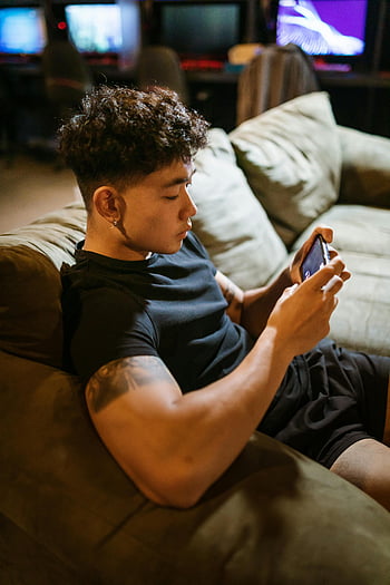 Man in Black Crew Neck T-shirt, Mobiel spel spelen, jonge man, Intens gefocust, gamen binnenshuis, smartphone, binnengamen, Casual gaming, mobiel gamen, divan, getatoeëerde arm