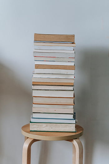 stack of books, wooden stool, plain background, vertical stack, neat pile, book stack, wooden surface, minimalist background, reading material, study resources, home decor