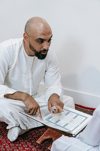 Mies valkoisessa thobessa, istuu maassa, Koraanin lukeminen, uskonnollinen teksti, puinen jalusta, sisäympäristö, perinteiset vaatteet, Islamilainen käytäntö, keskittynyt lukeminen, kulttuurinen puku, henkinen toiminta