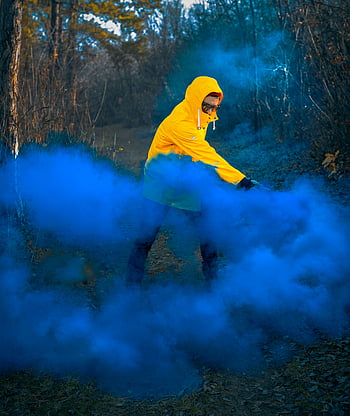 man met capuchon, blauwe rook, gele jas, bosrijke omgeving, dramatisch effect, blauwe rookbom, buitenfotografie, natuur scène, man in het bos, rook fotografie, dramatische verlichting