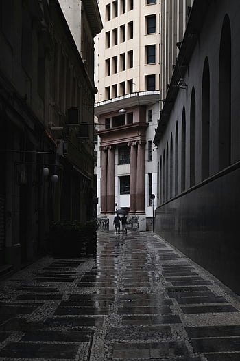 man walking, urban alley, wet pavement, reflections, towering architecture, dark cityscape, rainy day, city streets, architectural details, urban environment, street photography