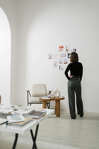 Wanita dengan lengan panjang hitam, celana abu -abu, kolase foto, kamar modern, dekorasi minimalis, suasana kreatif, gambar dinding, Melihat foto, interior putih, meja kayu, kursi putih