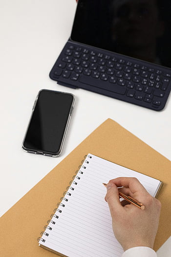 person writing, blank page, notebook, high-angle view, modern desk, smartphone, tablet keyboard, notepad, hand writing, workspace, productivity