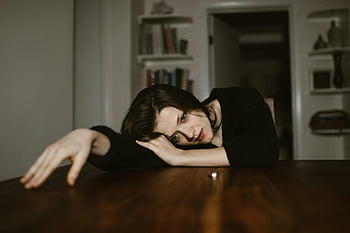 donna triste, tavolo di legno, stanza debolmente illuminata, umore riflessivo, donna premurosa, testa di riposo, Scena indoor, Espressione emotiva, Posa contemplativa, impostazione domestica, Background di libreria