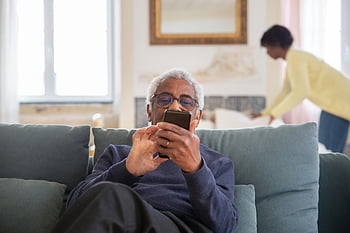 vanhempi mies, istuu sohvalla, älypuhelimen käyttäminen, kodikas kotiympäristö, sisä-, olohuone, rento asu, mukava asetus, rento asento, moderni teknologia, vanhusten elämäntapa