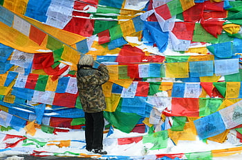 osoba, zelená béžová maskáčová bunda, tibetské modlitební praporky, zasněžené pozadí, věšení vlajek, zářivé barvy, zimní scény, venkovní aktivita, kulturní tradice, zasněžená zem, instalace modlitebních vlajek