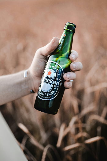 beer bottle, hand holding beer, outdoor field, lifestyle themes, beverage themes, green beer bottle, Heineken beer, malt lager, outdoor setting, casual drink, refreshing drink