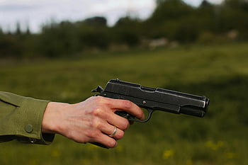hand holding gun, black handgun, silver ring, outdoor setting, close-up photo, pistol, firearm, outdoor firearm, hand with ring, green background, person holding gun