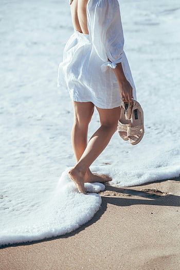 barefoot woman, white clothes, foamy sea, seaside stroll, holding sandals, beach walk, ocean waves, summer vacation, coastal scene, relaxed walk, beach fashion