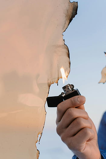 burning plastic, hand holding lighter, igniting paper, visible burn marks, flames, burning paper, lighter flame, plastic fire, fire ignition, outdoor burning, hand with lighter