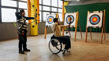 man in wheelchair, archery, indoor archery, focus, determination, adaptive sports, target practice, two adults, wheelchair user, archery targets, indoor sports