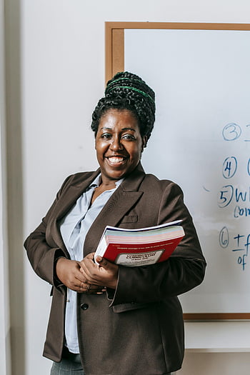 black female teacher, smiling lecturer, holding books, classroom whiteboard, education, teaching, professional woman, African American educator, workbooks, front of classroom, happy teacher