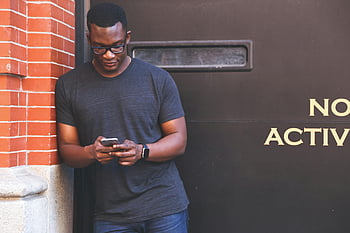 αφρικανικός αμερικανός άνδρας, smartphone, τοίχος από τούβλα, casual ενδυμασία, γκρι μπλουζάκι λαιμού πληρώματος, κλίση, ΕΞΩΤΕΡΙΚΟΥ ΧΩΡΟΥ, χρήση τεχνολογίας, αστικό περιβάλλον, ΤΡΟΠΟΣ ΖΩΗΣ, νέος ενήλικας