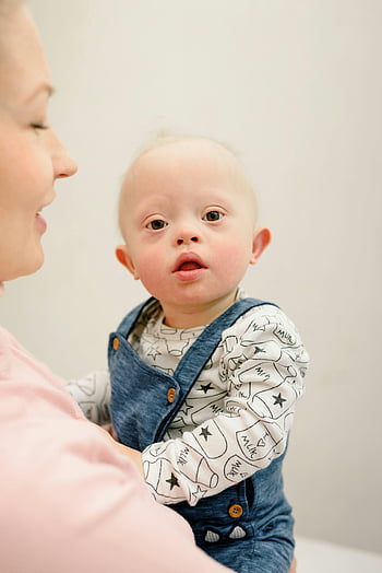woman carrying baby, adorable baby, Down syndrome, blue overalls, held by adult, indoors, baby with Down syndrome, blue denim overalls, adult holding baby, indoor photo, cute baby
