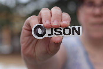JSON logo, hand holding sticker, close-up photo, outdoor setting, blurred background, JSON sticker, person holding logo, JSON symbol, JSON emblem, JSON design, JSON branding