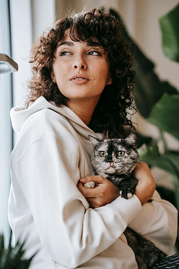 kvinne som holder katten, Koselig leilighet, hettegenser, Nær vindu, sjarmerende kvinne, innendørs foto, Katt på hendene, avslappet innstilling, hjemmemiljø, kjæledyr eier, Uformelt antrekk