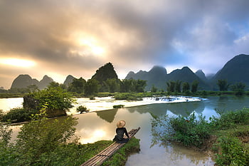 person sitting on bamboo, body of water, serene riverside, bamboo boat, sunset, lush greenery, natural landscape, calm waters, rural scene, peaceful setting, traditional boat