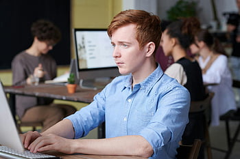 man using computer, young professional, working intently, busy modern office, colleagues around, laptop, desktop computer, office setting, focused work, professional attire, red-haired man