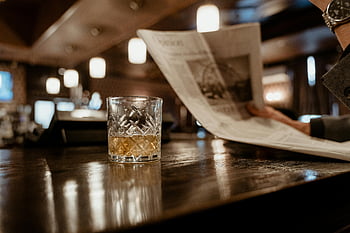 sklenice na whisky, detailní fotografie, barový pult, člověk čtení novin, Pozadí, matné osvětlení, křišťálové sklo, Amber kapalina, Vnitřní nastavení, Útulná atmosféra, Čtenář novin