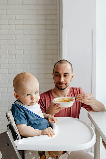 baby in blue shirt, white high chair, father feeding baby, toddler in high chair, brick wall background, family meal time, baby food, father and child, indoor family scene, baby bib, white highchair